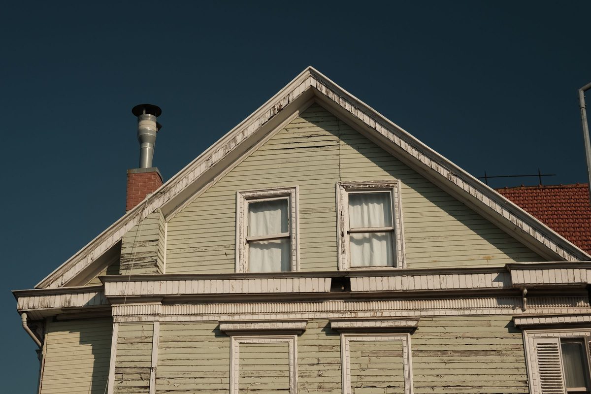 Weathered wooden gable with peeling trim and an open gap in the board below the attic windows