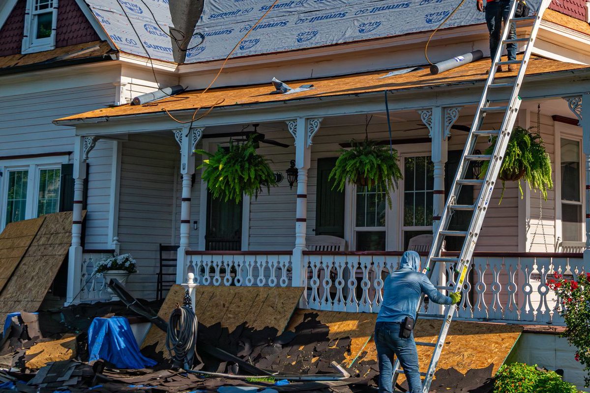 Worker steadying an extension ladder against the porch roof of an older house with a wraparound porch